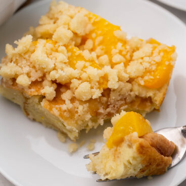 Close up of a slice of peach cobbler French toast on a white plate with a fork having taken a forkful of it.