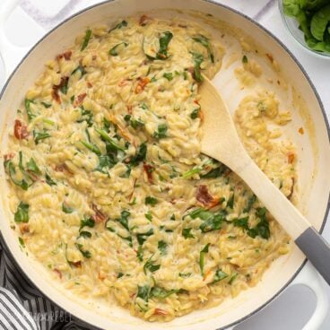 close up of a wooden ladle scooping sun dried tomato spinach orzo out of dish.